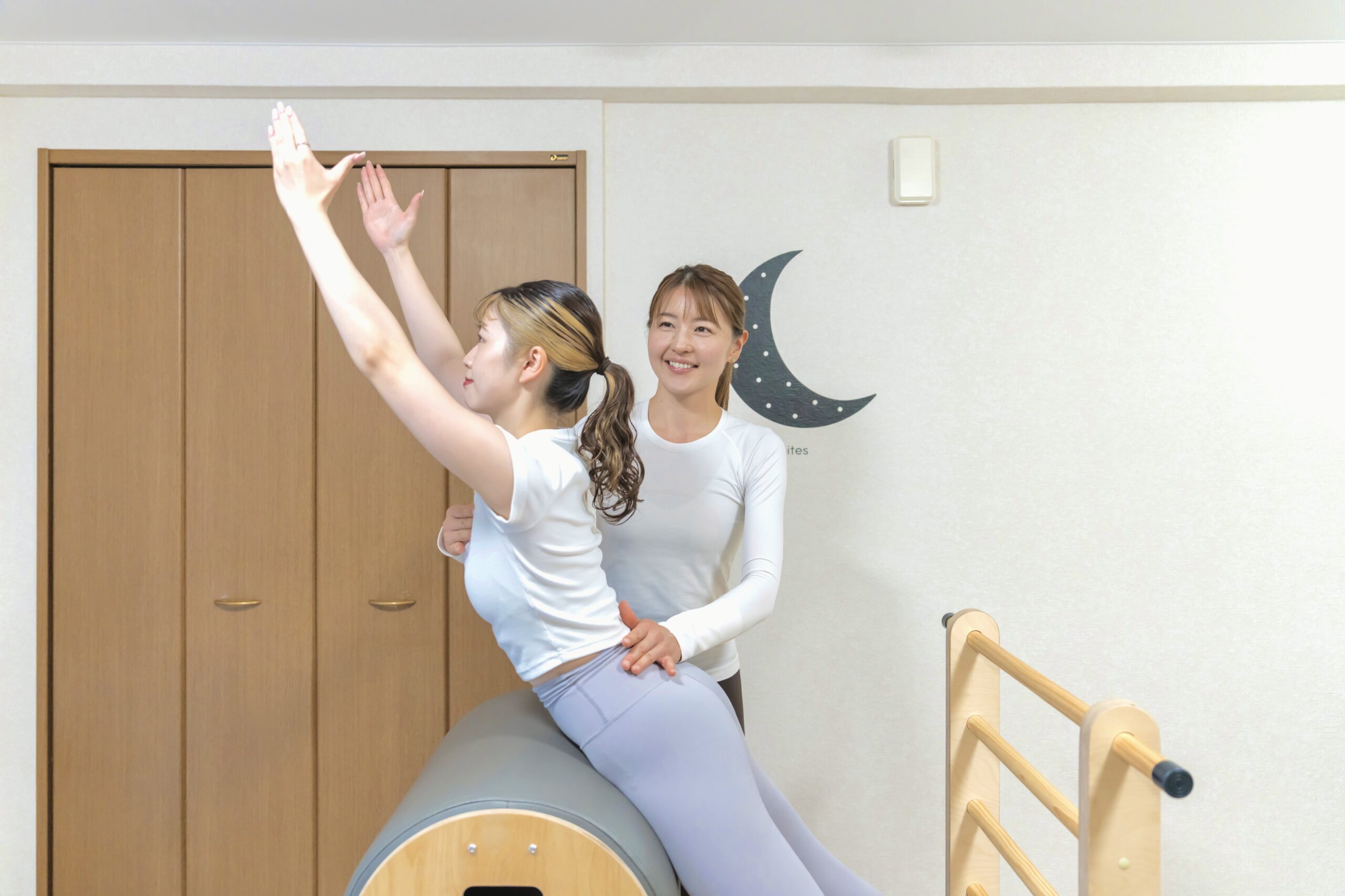 ピラティスバレルに体を預けて腕を上げる女性と、そのフォームをチェックするインストラクター