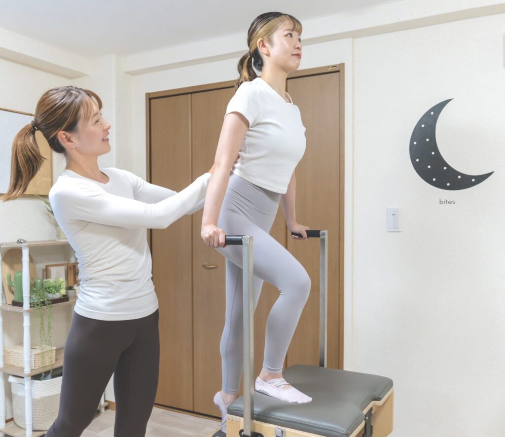 ピラティスマシンのバーを使って背筋を伸ばすポーズを行う女性と、それをサポートするインストラクター