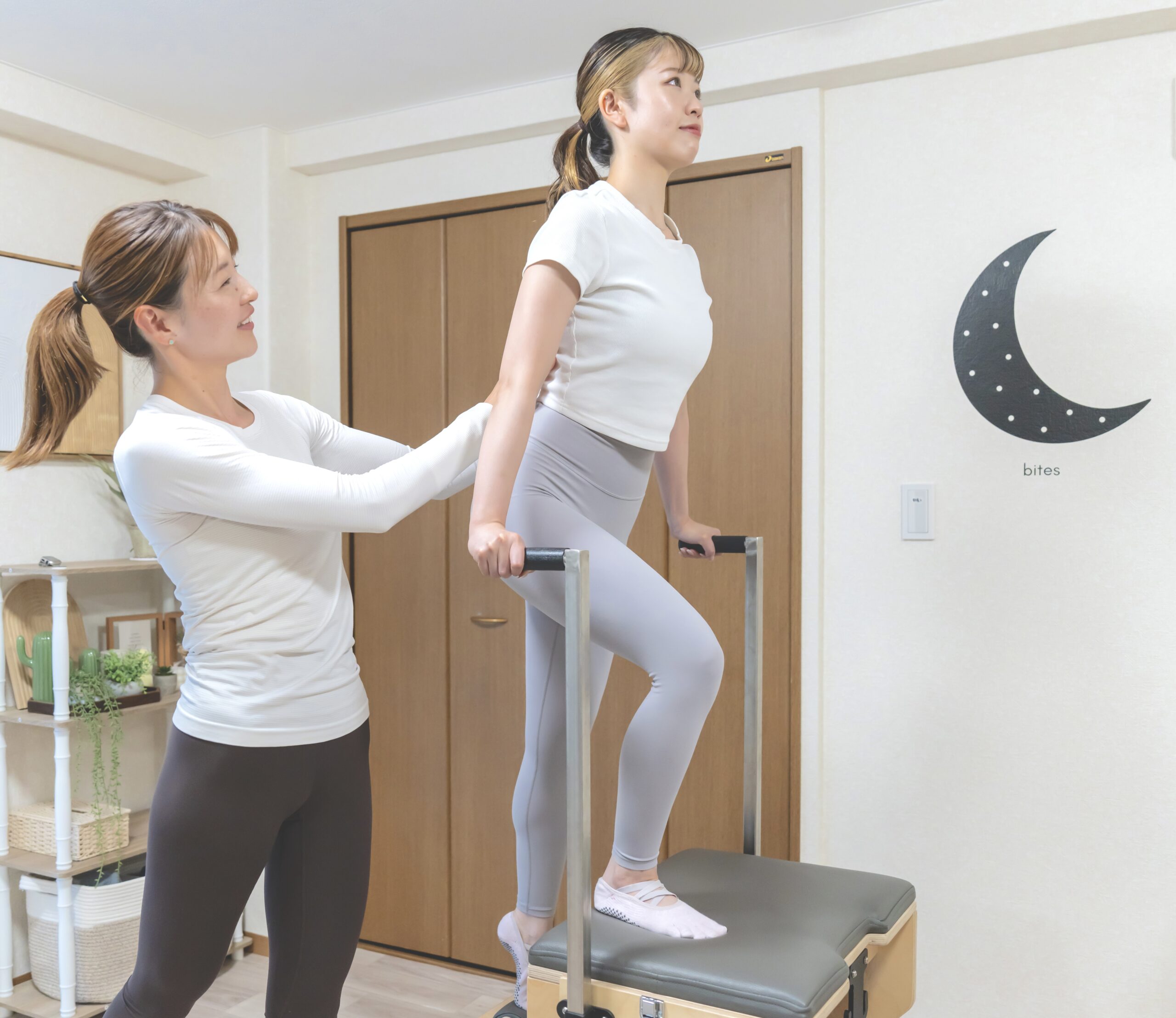 ピラティスマシンのバーを使って背筋を伸ばすポーズを行う女性と、それをサポートするインストラクター